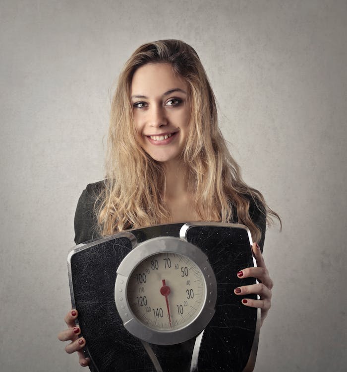 about-img Young woman with blond hair smiling, holding a vintage bathroom scale indoors.